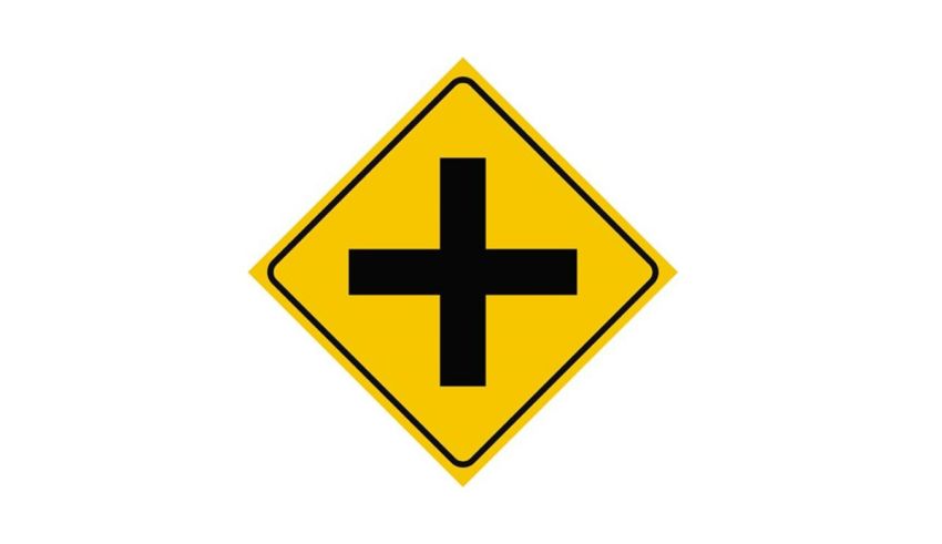 A yellow road sign with a black cross on it.