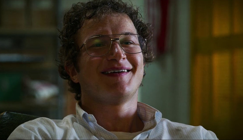 A man with glasses is smiling while sitting in a chair.