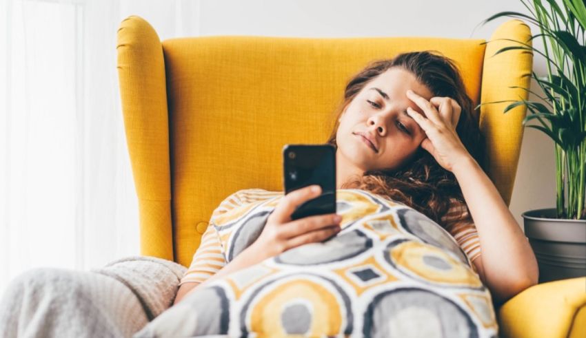 A pregnant woman looking at her phone while sitting in a yellow chair.