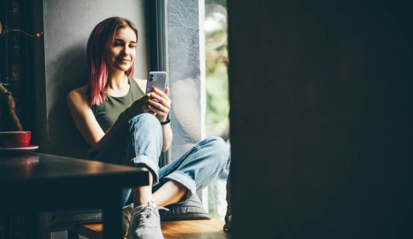 A young woman sitting on a window sill looking at her phone.