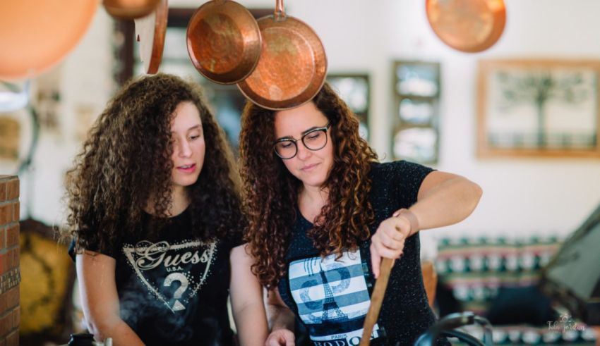Two women cooking in a kitchen with copper pots hanging above them.