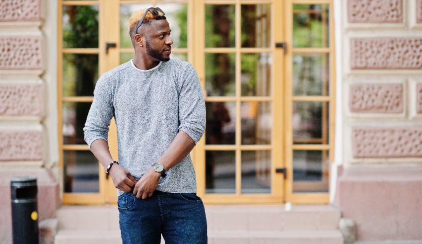 A young black man in a gray sweater and jeans standing in front of a building.