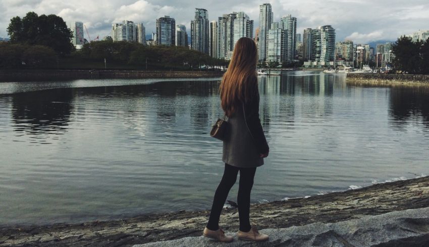 A woman is standing on a rock in front of a body of water with a city skyline in the background.