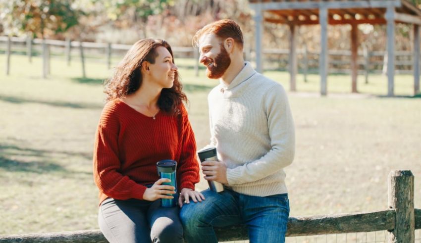 A couple sitting on a fence and drinking coffee.