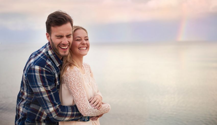 A man and woman hugging on the beach with a rainbow in the background.