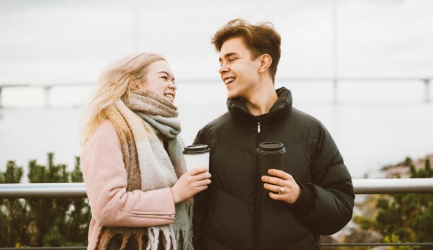 A young couple holding coffee cups in front of a bridge.