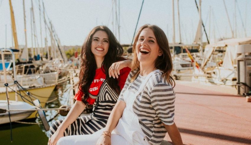 Two young women sitting on a dock in a marina.