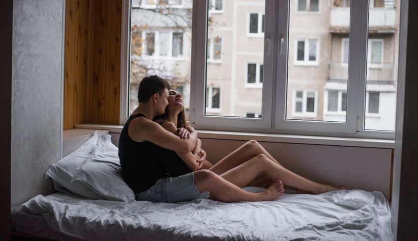 A man and woman sitting on a bed in front of a window.