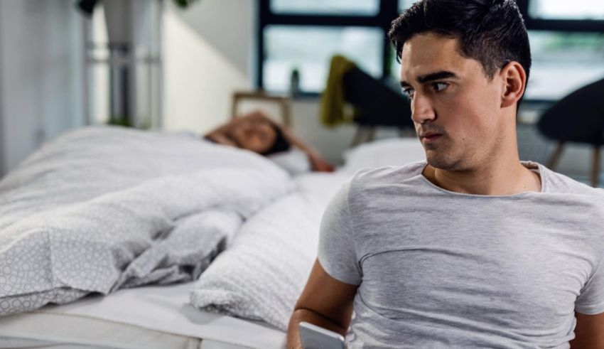 A man looking at his phone while laying in bed with another man.