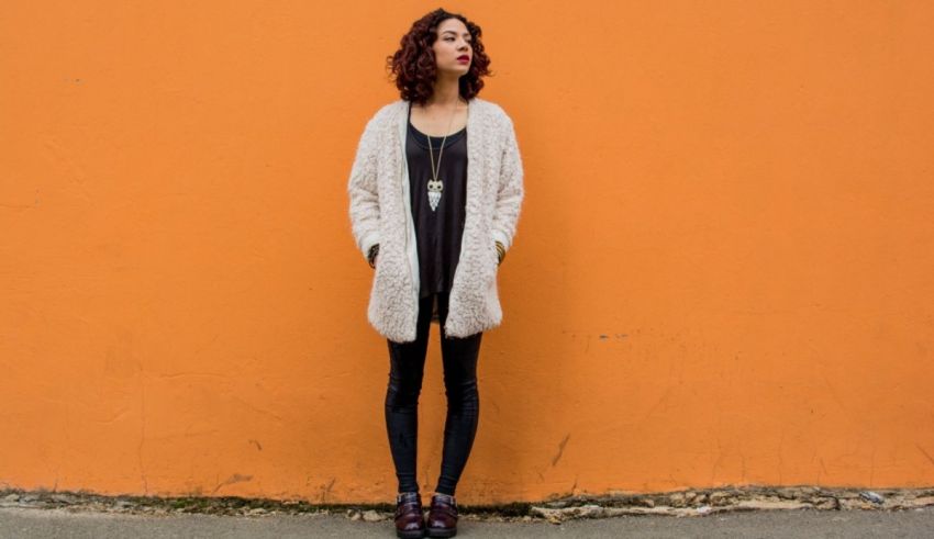 A woman wearing a white cardigan and black leggings in front of an orange wall.