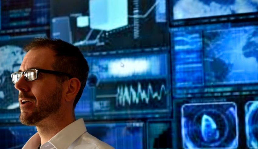 A man in glasses standing in front of a computer screen.