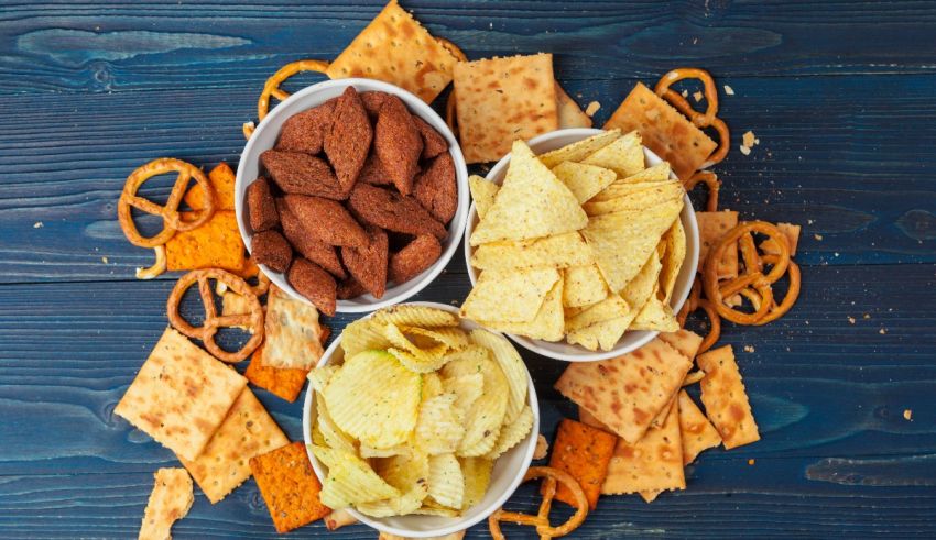 Three bowls of chips, crackers and pretzels on a blue background.