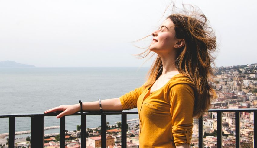 A woman in a yellow shirt is standing on a balcony overlooking the sea.