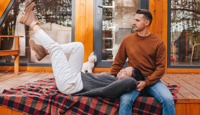A man and woman sitting on a blanket in front of a wooden house.