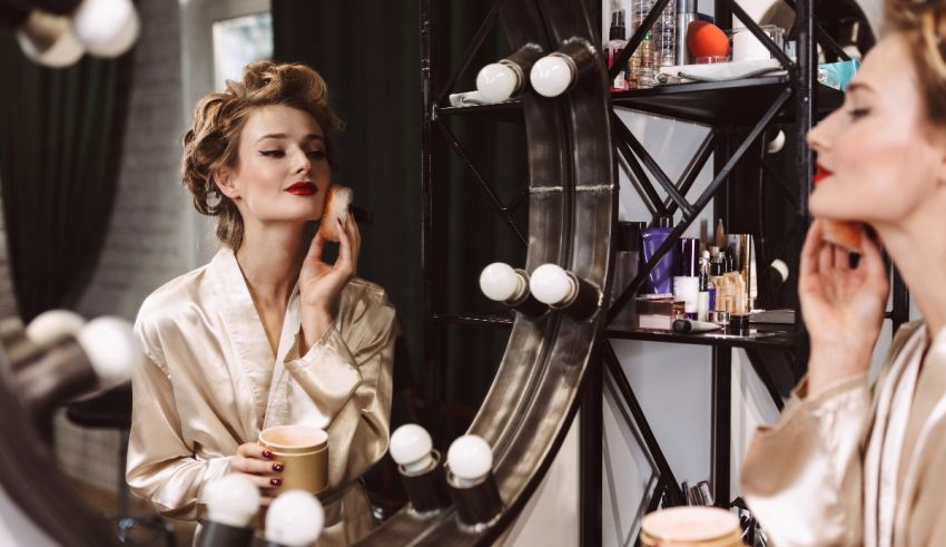 A woman in a robe is applying makeup in front of a mirror.
